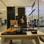 Innenansicht eines Luxusgeschäfts mit einem Ausstellungstisch mit Lederwaren, darunter Handtaschen und Brieftaschen. Hinter dem Tisch befindet sich ein Bildschirm, auf dem ein Modell zu sehen ist. Das Geschäft ist gut beleuchtet und im Hintergrund sind Kleidung und Brillen zu sehen.