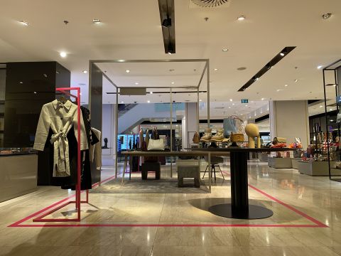 Ein modernes Ladeninterieur, in dem elegante Kleidung und Accessoires präsentiert werden. Auf einem zentralen Tisch werden verschiedene Taschen, Hüte und Kleinigkeiten ausgestellt. Schaufensterpuppen auf der linken Seite präsentieren stilvolle Outfits. Der Raum ist gut beleuchtet und minimalistisch gestaltet.