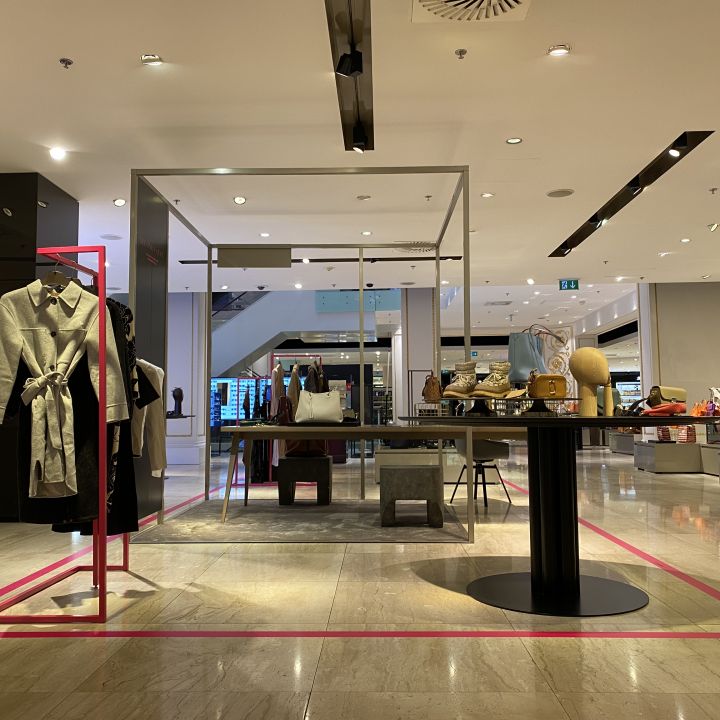 Ein modernes Ladeninterieur, in dem elegante Kleidung und Accessoires präsentiert werden. Auf einem zentralen Tisch werden verschiedene Taschen, Hüte und Kleinigkeiten ausgestellt. Schaufensterpuppen auf der linken Seite präsentieren stilvolle Outfits. Der Raum ist gut beleuchtet und minimalistisch gestaltet.