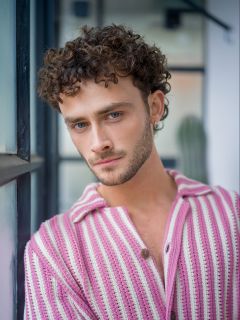 Ein junger Mann mit lockigen braunen Haaren und einem kurzen Bart trägt einen rosa-weiß gestreiften Pullover, steht drinnen an einem Fenster und schaut in die Kamera.