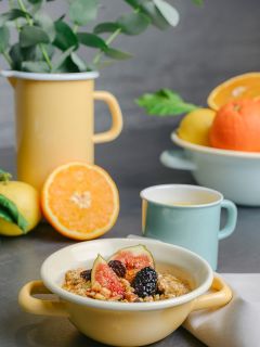 Eine Schüssel Haferflocken mit Feigen und Brombeeren, daneben ein Becher, Orangen, Zitronen und ein gelber Krug mit grünen Blättern im Hintergrund.