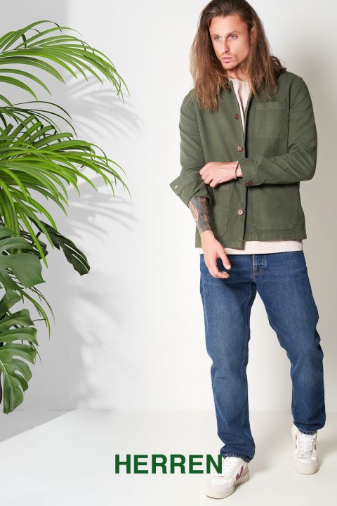 Ein Mann mit langen Haaren trägt eine olivgrüne Jacke, ein weißes Hemd, blaue Jeans und weiße Turnschuhe. Er steht neben einer Blattpflanze vor einem schlichten hellen Hintergrund. Unten ist das Wort „HERREN“ zu sehen.
