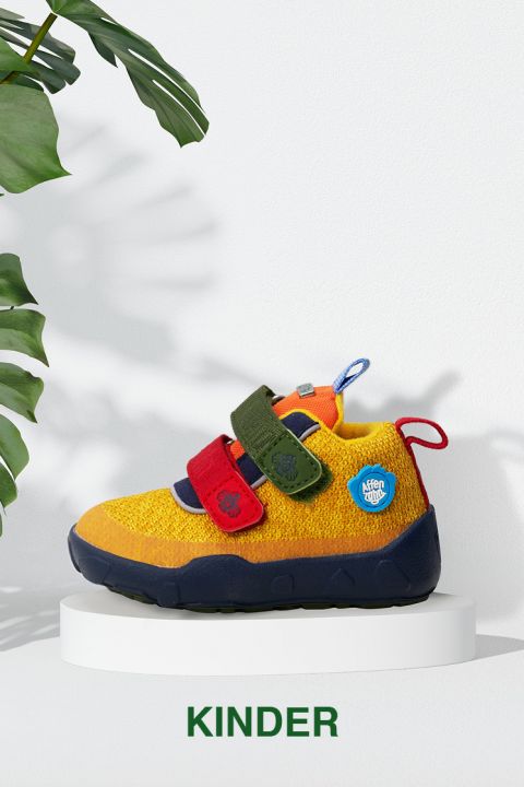 Ein farbenfroher Kinderschuh mit blauen, gelben und roten Abschnitten auf einem weißen runden Ständer. Der Schuh hat zwei Klettverschlüsse – rot und grün – und dekorative Aufnäher. Er ist vor einem weißen Hintergrund mit grünen Blättern auf der linken Seite zu sehen. Der Text lautet „KINDER.