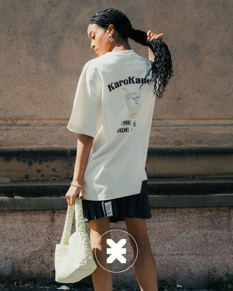 Eine Frau mit einem langen geflochtenen Pferdeschwanz steht im Freien und blickt abgewandt. Sie trägt ein cremefarbenes T-Shirt mit Grafik, einen schwarzen Rock und hält eine gesteppte hellgelbe Handtasche in der Hand. Sonnenlicht hebt ihr Outfit vor einer strukturierten Wand hervor.