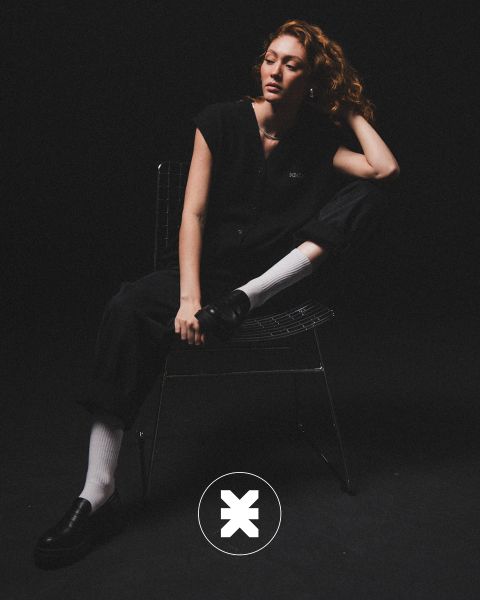 Eine Person mit lockigem Haar sitzt auf einem Drahtstuhl in einem dunklen Raum, bekleidet mit schwarzer Kleidung, weißen Socken und schwarzen Halbschuhen. Ein Bein ist angewinkelt, ein Ellbogen liegt auf dem Knie, und auf dem Boden befindet sich ein kreisförmiges Logo mit einem "X".