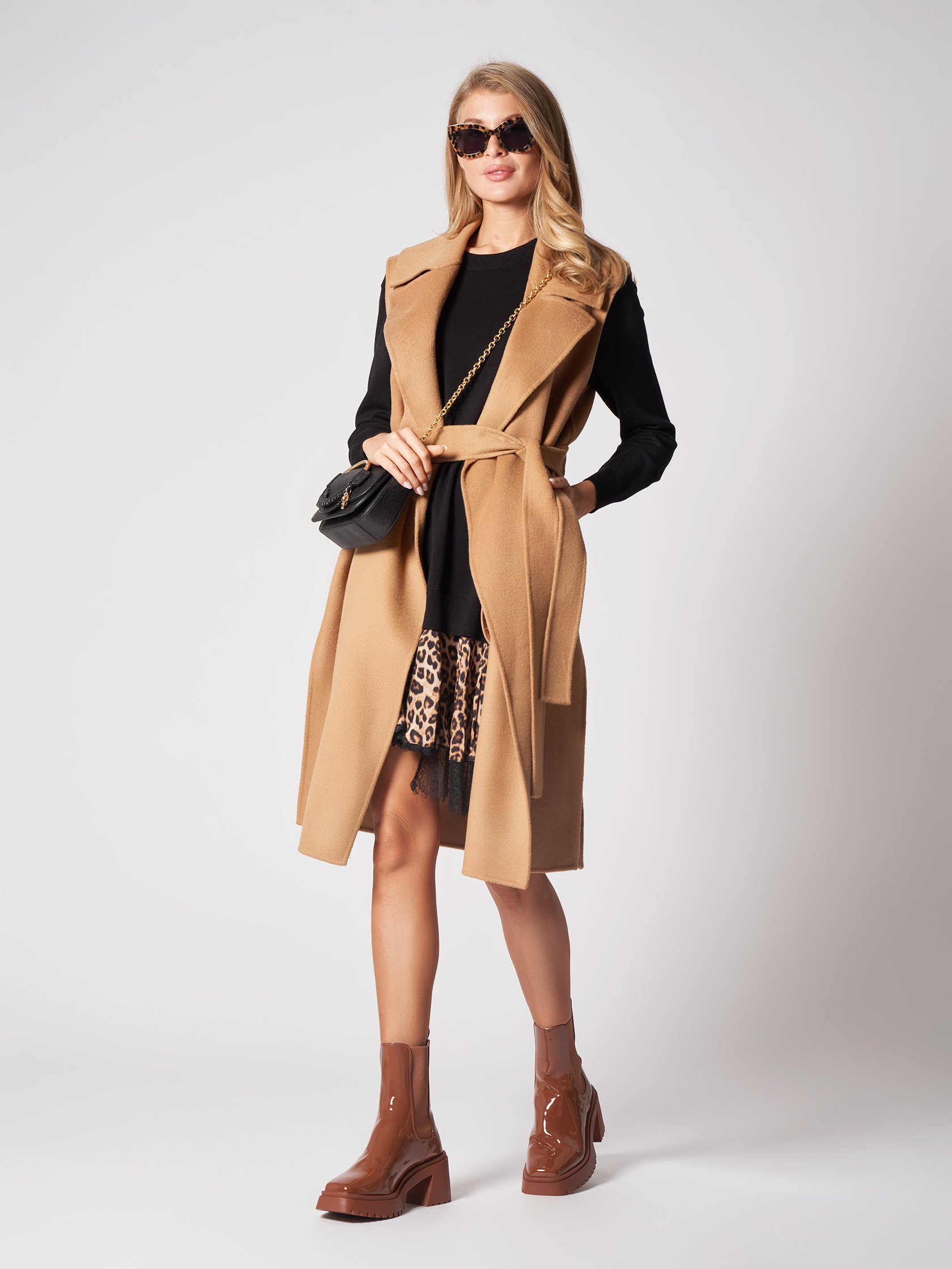 Eine Person trägt ein schwarzes Kleid mit einem beigen ärmellosen Mantel, einem Rock mit Leopardenmuster und braunen Stiefeln. Sie hat lange blonde Haare, eine Sonnenbrille und eine schwarze Handtasche mit Kettenriemen und steht vor einem schlichten hellen Hintergrund.