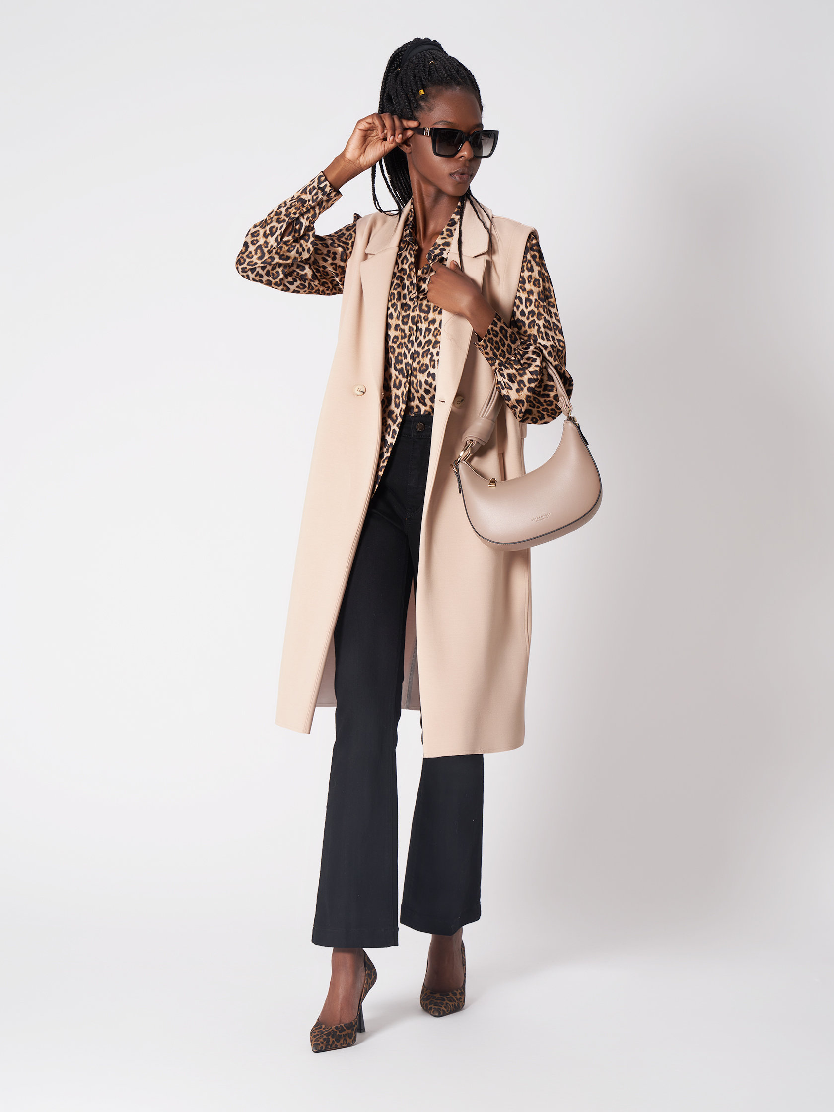 Eine stylische Person mit Sonnenbrille, Bluse mit Leopardenmuster, beigem ärmellosem Mantel, schwarzen Hosen und High Heels mit Leopardenmuster posiert, wobei sie mit einer Hand die Sonnenbrille zurechtrückt und mit der anderen eine beige Handtasche vor einem schlichten Hintergrund hält.