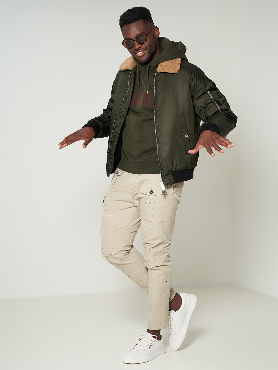 Ein Mann mit Sonnenbrille, grünem Kapuzenpullover und olivfarbener Bomberjacke posiert selbstbewusst. Er trägt hellbeige Cargohosen und weiße Sneakers. Der Hintergrund ist schlicht und hell gehalten.
