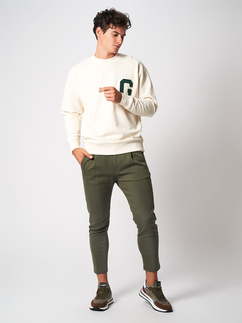 Eine Person, die einen cremefarbenen Pullover mit dem Buchstaben „G“ darauf und olivgrüne Hosen trägt, steht vor einem schlichten Hintergrund. Sie blickt zur Seite und hat eine Hand leicht erhoben. Sie trägt außerdem braune Turnschuhe.
