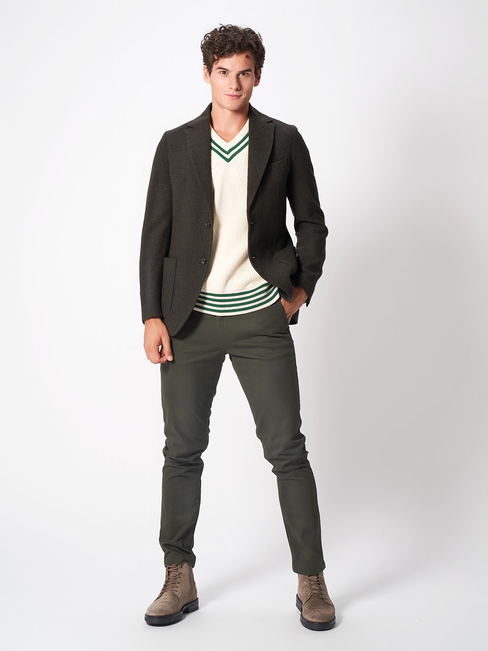 Eine Person mit lockigem Haar trägt einen dunklen Blazer über einem weiß-grünen Pullover mit V-Ausschnitt, dazu dunkelgrüne Hosen und hellbraune Stiefel. Sie steht vor einem schlichten weißen Hintergrund, lächelt leicht und hat die Hände in den Taschen.