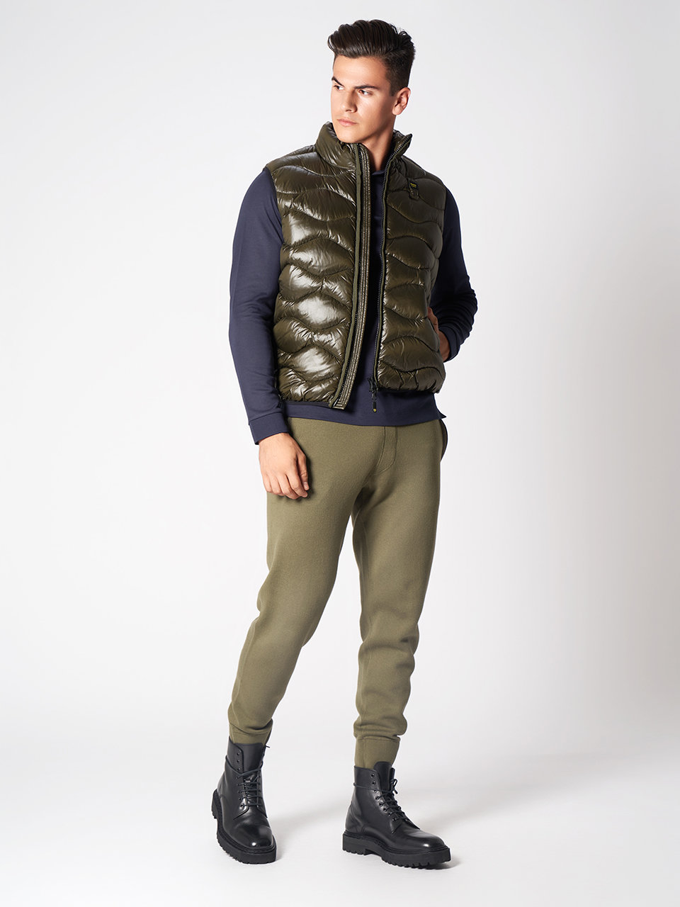Eine Person steht da und trägt eine glänzende olivgrüne Pufferweste über einem dunklen Langarmhemd, olivgrüne Jogginghosen und schwarze Stiefel. Sie hat kurze Haare und posiert vor einem schlichten, hellen Hintergrund.