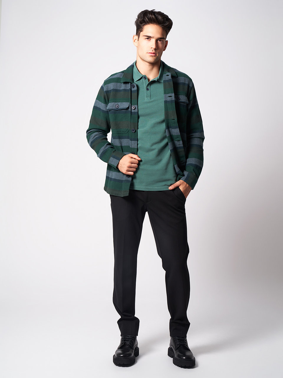 Ein Mann steht vor einem schlichten Hintergrund. Er trägt eine grün gestreifte Jacke über einem grünen Poloshirt, dazu eine schwarze Hose und schwarze Schuhe. Er blickt selbstbewusst in die Kamera, eine Hand in der Tasche.