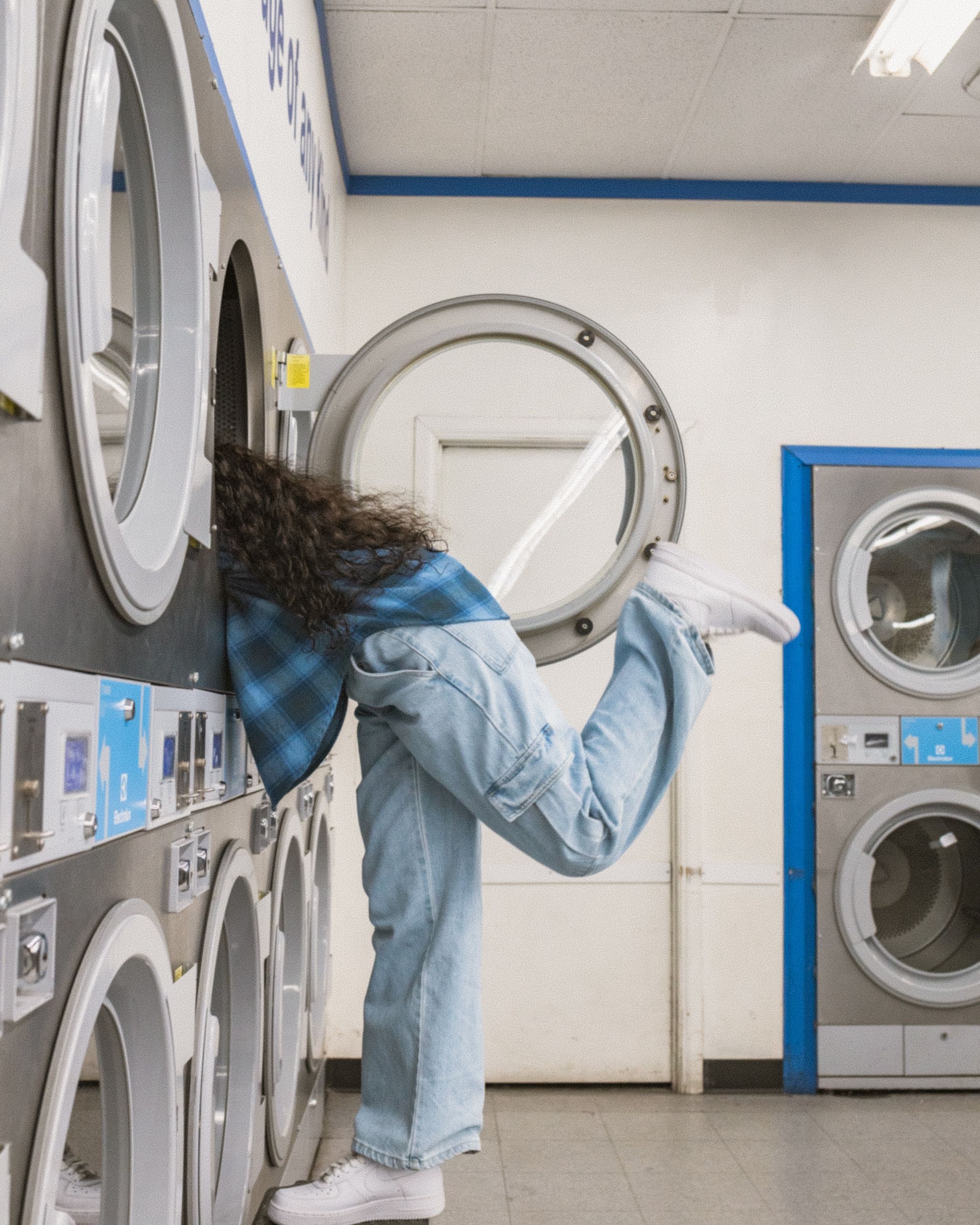 Eine Person in einem blau karierten Hemd und Jeans lehnt sich in einem Waschsalon an eine Waschmaschine. Ihr rechtes Bein ist zum Balancieren nach hinten angehoben. Der Raum verfügt über mehrere Waschmaschinen und Trockner in blau-weißen Farbtönen.