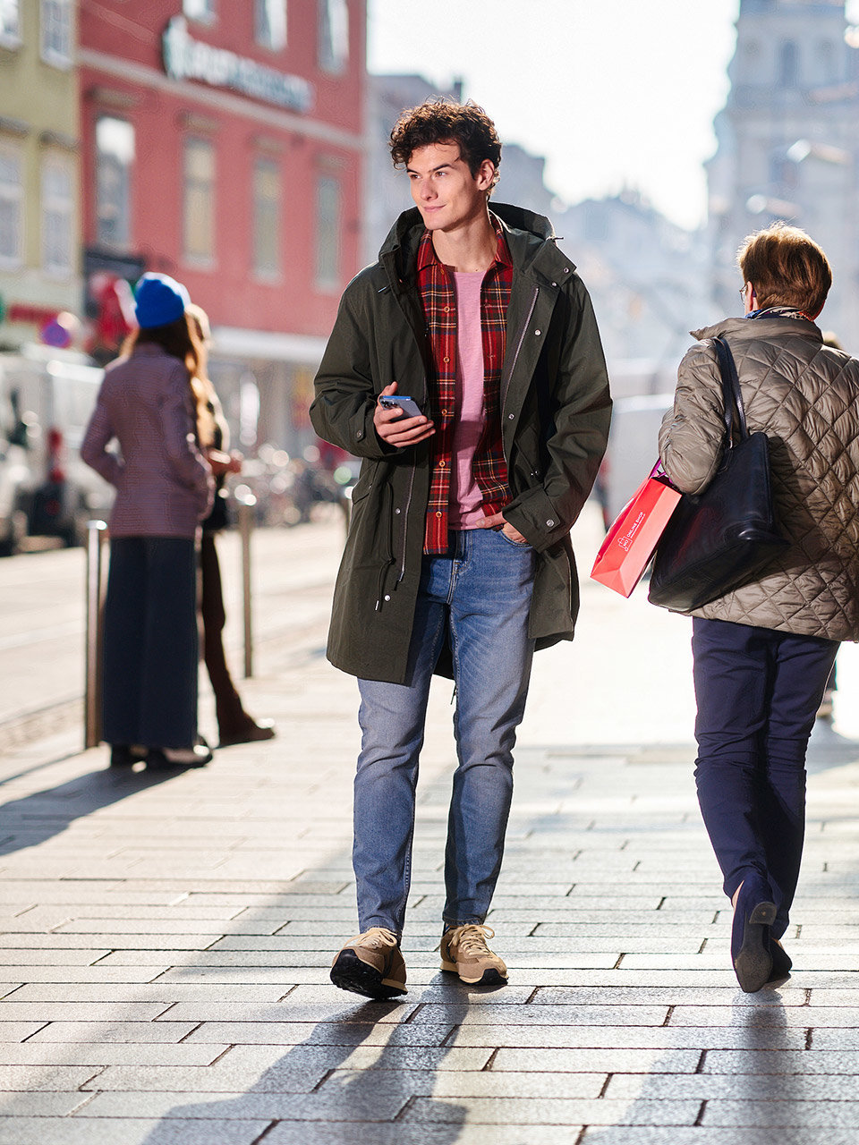 Ein junger Mensch läuft eine Stadtstraße entlang und schaut dabei auf sein Handy. Er trägt einen grünen Mantel und Jeans. Auf der Straße herrscht reger Verkehr, im Hintergrund säumen bunte Gebäude das Bild, während das Sonnenlicht lange Schatten wirft.