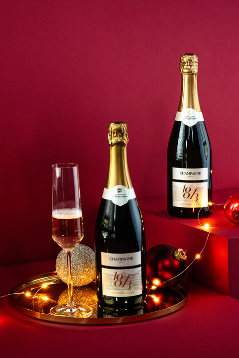 Zwei Champagnerflaschen mit goldenen Etiketten stehen auf einem goldenen Tablett vor einem satten roten Hintergrund. Ein gefülltes Champagnerglas, dekorative Ornamente und eine Lichterkette sorgen für eine festliche Note.