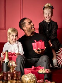 Ein Mann sitzt mit einer roten Geschenkbox auf einem roten Sofa und lächelt zwei kleine Kinder an. Ein Kind, weiß gekleidet, hält ein ähnliches rotes Geschenk, während das andere, schwarz gekleidet, auf der Sofalehne sitzt. Im Vordergrund steht ein Tisch mit Leckereien und einer Flasche.