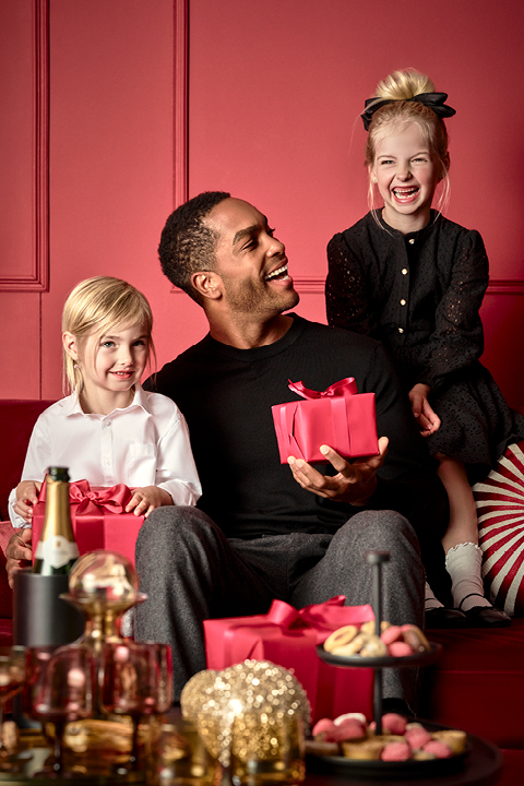 Ein Mann sitzt mit einer roten Geschenkbox auf einem roten Sofa und lächelt zwei kleine Kinder an. Ein Kind, weiß gekleidet, hält ein ähnliches rotes Geschenk, während das andere, schwarz gekleidet, auf der Sofalehne sitzt. Im Vordergrund steht ein Tisch mit Leckereien und einer Flasche.
