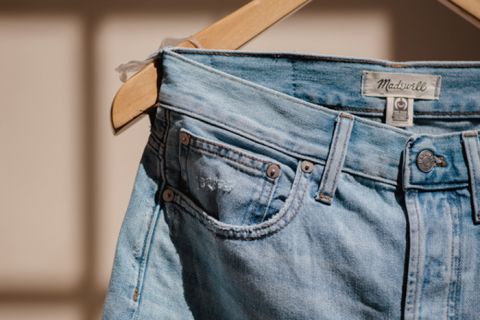 Nahaufnahme einer hellblauen Jeans, die auf einem hölzernen Kleiderbügel hängt. Die Jeans hat am Hosenbund ein Lederetikett mit dem Markennamen „Madewell“ und ist in warmes Sonnenlicht getaucht, wodurch ihre verblasste Textur und Nähte hervorgehoben werden.