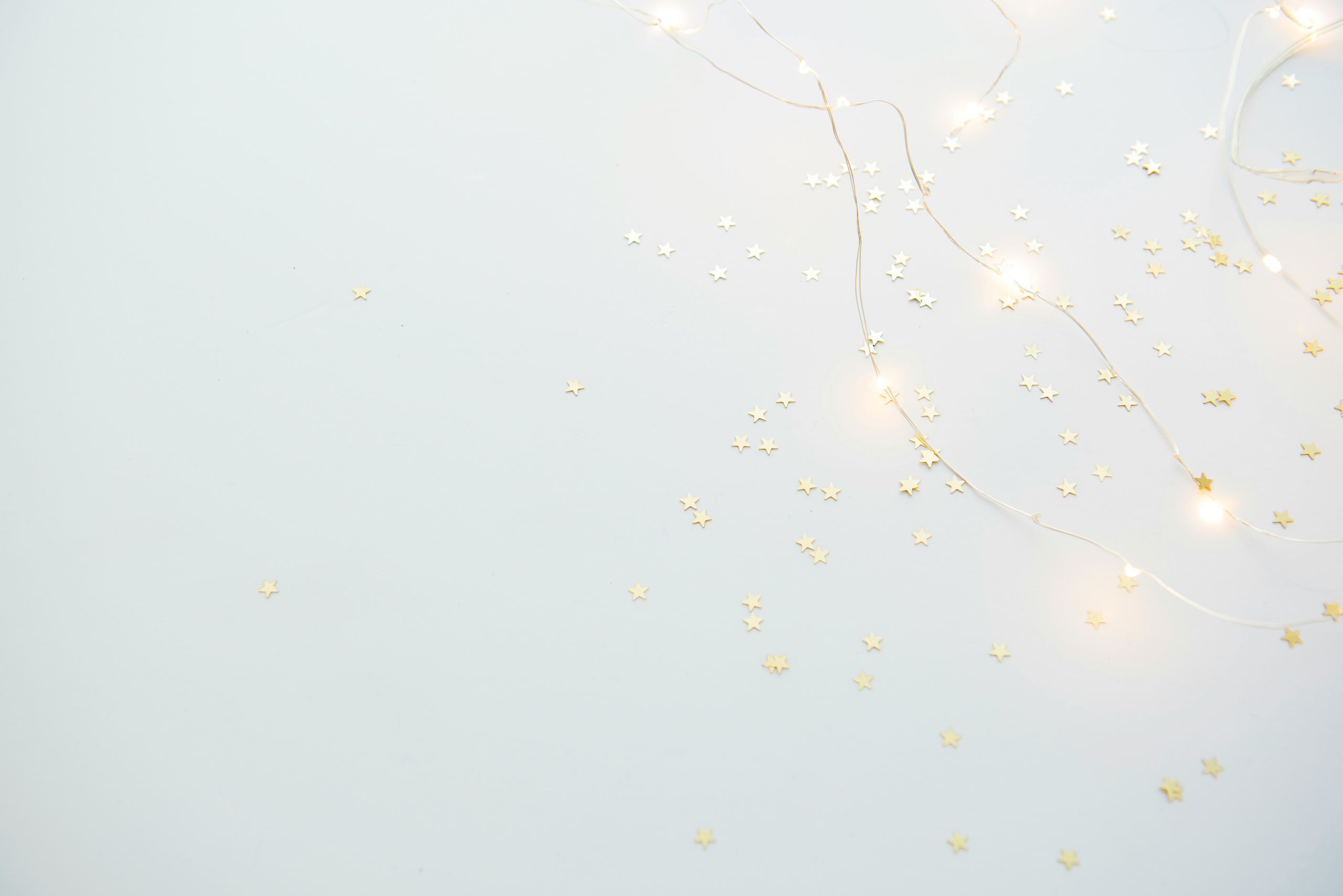 Ein hellgrauer Hintergrund mit kleinen goldenen Konfettisternen, die über die Oberfläche verstreut sind. Zwischen den Sternen sind auch zarte, leuchtende Lichterketten verflochten. Die Szene ist minimalistisch und festlich.