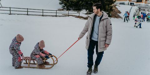 Ein Mann in einer Winterjacke zieht einen Holzschlitten mit zwei darauf sitzenden Kindern, die passende Schneeanzüge mit Leopardenmuster und rosa Mützen tragen, durch eine verschneite Landschaft. In der Ferne sieht man andere Menschen, die die verschneite Umgebung genießen.