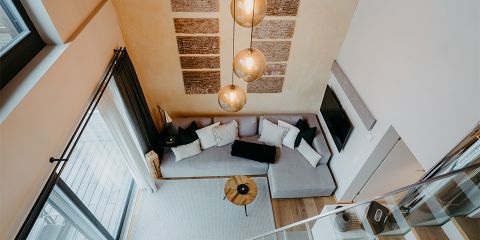 Ein modernes Wohnzimmer mit einem gemütlichen, L-förmigen, grauen Sofa, geschmückt mit schwarzen und weißen Kissen. In der Mitte steht ein runder Couchtisch aus Holz. Drei kunstvolle Hängelampen hängen herab, an der Wand dahinter hängen Kunstwerke. Große Fenster sorgen für natürliches Licht.