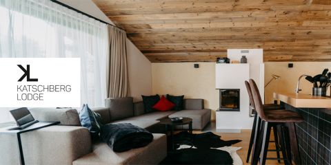 Ein gemütlicher Wohnbereich mit modernem Design, mit einem Ecksofa mit roten und grauen Kissen, einem Kamin, einem Kuhfellteppich und einer Küchenbar mit Hockern. Der Raum hat eine Holzdecke und große Fenstervorhänge. Der Logotext lautet „Katschberg Lodge“.