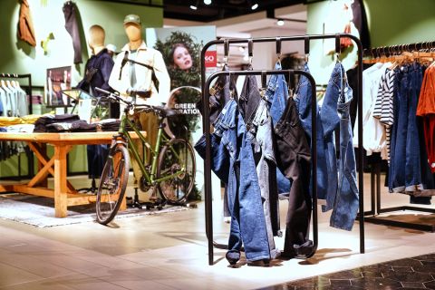 Innenansicht eines Bekleidungsgeschäfts mit verschiedenen Jeans, die im Vordergrund an einem Ständer hängen. Im Hintergrund sind ein Fahrrad und Schaufensterpuppen in Freizeitkleidung neben einem Tisch mit ordentlich gefalteter Kleidung ausgestellt. Die Kulisse ist modern eingerichtet.