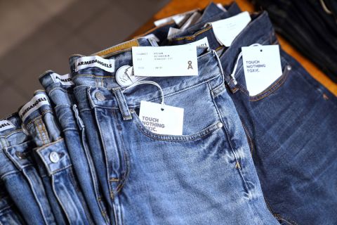 Eine Auslage ordentlich gefalteter Bluejeans in einem Geschäft. Am Hosenbund und im Taschenbereich sind Etiketten zu sehen. Die Jeans sind in verschiedenen Blautönen gehalten und mit Etiketten versehen, die auf umweltfreundliche oder ungiftige Materialien hinweisen.