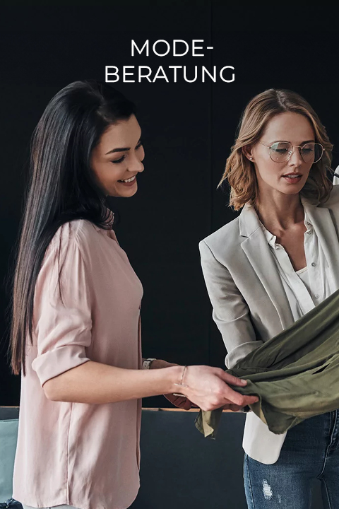 Zwei Frauen stehen in einem Raum mit dunkler Wand. Die eine hat lange dunkle Haare, trägt ein rosa Shirt und lächelt. Die andere hat blonde Haare und eine Brille, trägt einen hellen Blazer und hält ein grünes Kleidungsstück in der Hand. Über ihnen steht der Schriftzug „Mode-Beratung“.