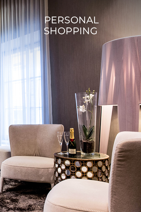 Luxuriöses Zimmer mit zwei beigen Stühlen, einem runden Tisch mit einer Champagnerflasche und Gläsern und einer Orchidee in einer Vase. Daneben steht eine große, moderne Lampe. Oben steht der Text „Personal Shopping“.