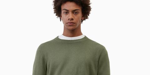 Eine Person mit lockigem Haar, die einen grünen Pullover über einem weißen Hemd trägt, blickt vor einem weißen Hintergrund direkt in die Kamera.