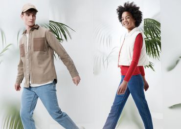 Ein Mann in beiger Jacke, blauen Jeans und beiger Mütze und eine Frau in weißer Weste, rotem Pullover und blauen Jeans gehen selbstbewusst in einer hellen, stilvollen Umgebung mit großen grünen Blättern im Hintergrund.