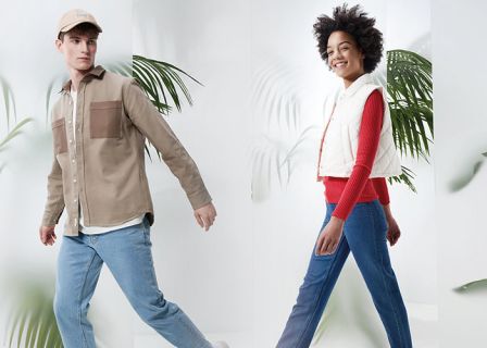 Ein Mann in beiger Jacke, blauen Jeans und beiger Mütze und eine Frau in weißer Weste, rotem Pullover und blauen Jeans gehen selbstbewusst in einer hellen, stilvollen Umgebung mit großen grünen Blättern im Hintergrund.