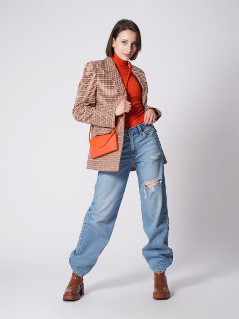 Eine Person posiert selbstbewusst vor einem schlichten Hintergrund. Sie trägt einen karierten Blazer, einen orangefarbenen Rollkragenpullover, Jeans im Used-Look und braune Stiefel. Eine orangefarbene Umhängetasche sorgt für einen Farbtupfer. Die Pose ist lässig und doch stilvoll.