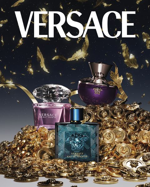 Flakons von Versace-Parfums – Bright Crystal, Dylan Purple und Eros Eau de Toilette – sind von einem Haufen Goldmünzen und Federn umgeben. Das Versace-Logo ist oben auf einem dunklen Hintergrund deutlich zu sehen.