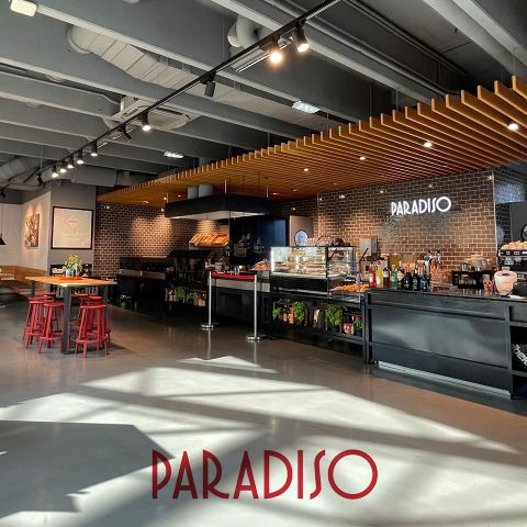 Modernes Restaurant-Interieur mit elegantem Design. Schwarze Theke mit verschiedenen Artikeln und „PARADISO“-Schild an der Wand. Holzakzente an der Decke und rote Holzhocker neben hohen Tischen. Sonnenlicht fällt durch große Fenster auf den polierten grauen Boden.
