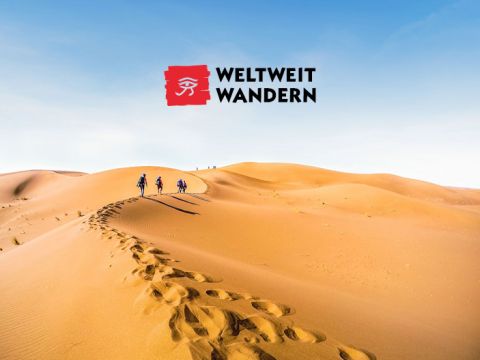 Eine Gruppe von Menschen wandert unter einem klaren blauen Himmel auf Sanddünen. Fußspuren hinterlassen Spuren. Das Logo „Weltweit Wandern“ ist im Himmelsbereich des Bildes deutlich zu sehen.