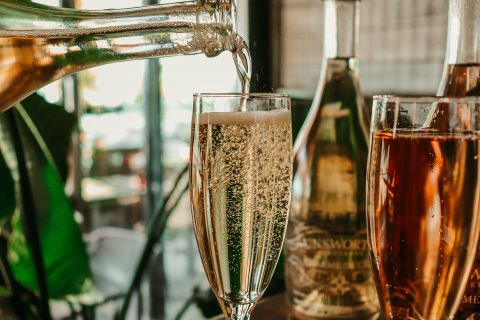 Nahaufnahme eines Glases, das mit Sekt aus einer Flasche gefüllt wird. Im Hintergrund sind zwei weitere Flaschen zu sehen, eine mit einem rosafarbenen Getränk. Im Glas steigen sichtbar Bläschen auf, vor einem verschwommenen, gemütlichen Innenraum mit viel Grün.