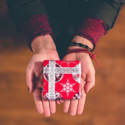Hände halten ein kleines, festlich verpacktes Geschenk mit rotem Papier und silberner Schleife.