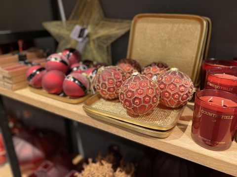 Eine Auswahl festlicher rot-goldener Weihnachtsdekorationen und roter Kerzen auf einem Holzregal. Die Dekorationen gibt es in zwei Stilen: glattes Rot mit Streifen und strukturiertes Rot mit kreisförmigen Mustern. Ein großer Stern schmückt den Hintergrund.