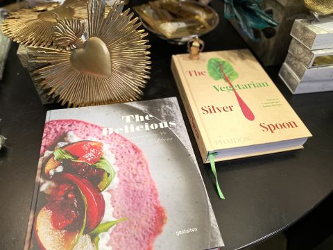 Zwei Kochbücher auf einem Tisch: „The Delicious“ mit einer Nahaufnahme eines bunten Gerichts auf dem Cover und „The Vegetarian Silver Spoon“ mit minimalistischem Design und einem Karottenbild. In der Nähe befinden sich dekorative Gegenstände, darunter ein goldenes Herzornament.