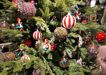 Ein Weihnachtsbaum, der mit bunten Ornamenten geschmückt ist, darunter große rot-weiß gestreifte Kugeln und Miniaturfiguren. Der Baum ist mit üppigen grünen Zweigen behangen und sorgt für eine festliche und lebendige Weihnachtsstimmung.