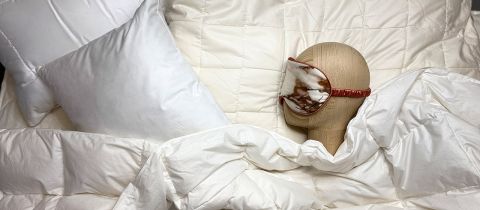 Ein hölzerner Puppenkopf trägt eine gemusterte Schlafmaske und liegt auf einem Bett mit weißen Kissen und einer gesteppten Bettdecke. Die Szene vermittelt eine gemütliche und erholsame Atmosphäre.