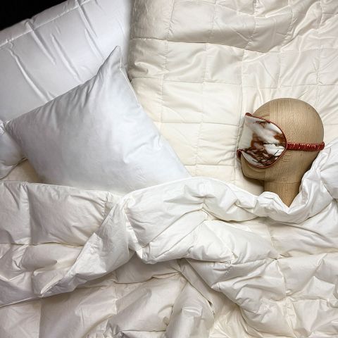 Eine gemütliche Schlafzimmerszene mit einem weißen Kissen und einer Bettdecke. Ein Puppenkopf mit einer Schlafmaske mit Marmormuster ruht auf dem Bett, umgeben von der flauschigen Bettwäsche.