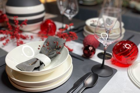 Eine festliche Tischdekoration mit gestapelten weißen Tellern, einer grauen Serviette mit roten Beeren, schwarzem Besteck und Weingläsern. Dekorative rote Kugeln und Beerenzweige betonen das elegante Arrangement.