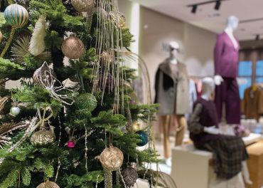 Im Vordergrund steht ein geschmückter Weihnachtsbaum mit goldenen und grünen Ornamenten. Im Hintergrund präsentieren Schaufensterpuppen Winteroutfits in einem Bekleidungsgeschäft.