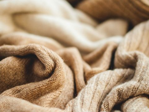 Nahaufnahme eines Stapels aus weichem, gestricktem Wollstoff in verschiedenen Beige- und Brauntönen. Die Texturen sind unterschiedlich, wobei einige Bereiche leicht gerippt erscheinen. Die Beleuchtung hebt die sanften Falten und das weiche Material hervor.