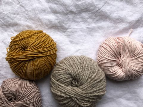 Vier Garnknäuel in Senfgelb, Beige, Grau und Hellrosa sind auf einem strukturierten weißen Stoffhintergrund angeordnet.