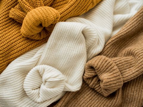 Eine Sammlung gemütlicher Strickpullover in Senfgelb-, Creme- und Brauntönen ist ordentlich arrangiert. Die Ärmel jedes Pullovers sind zu einem Knoten zusammengebunden, wodurch die Textur und Wärme des Strickstoffs zur Geltung kommt.