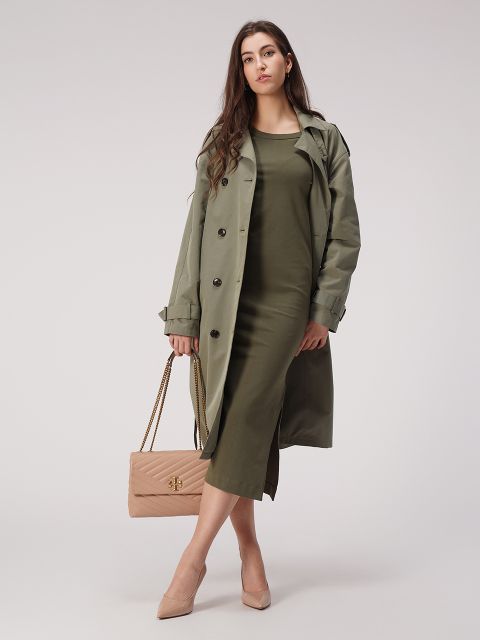 Eine Frau in einem olivgrünen Kleid und einem dazu passenden Trenchcoat hält eine beige gesteppte Handtasche mit Kettenriemen. Sie trägt beige High Heels und steht vor einem schlichten grauen Hintergrund und blickt selbstbewusst in die Kamera.